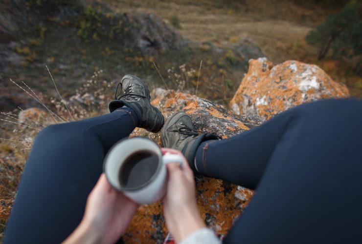 Hands of a girl with cup of coffee and trekking boots on backgro